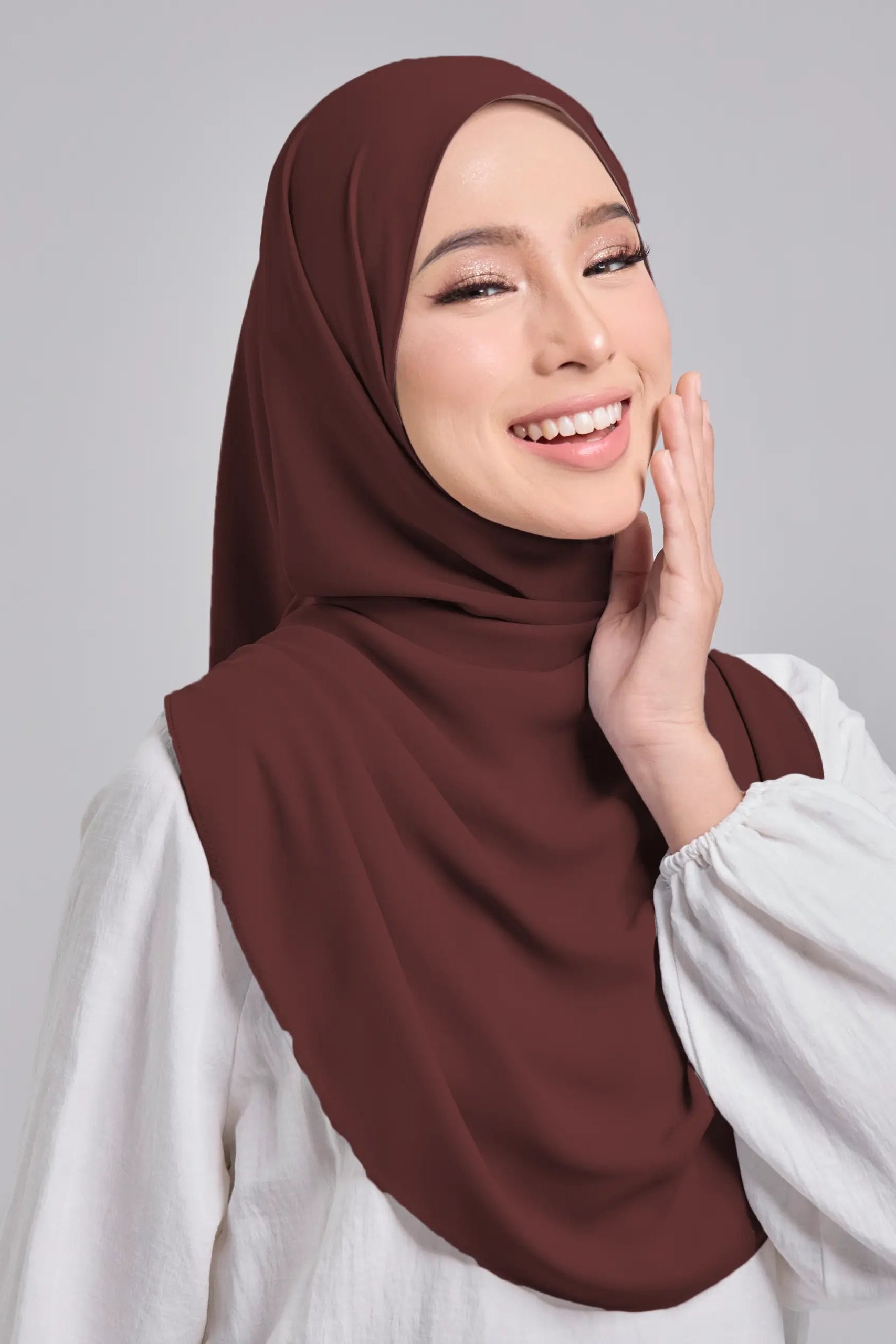 Woman wearing a brown hijab and white shirt against a gray background