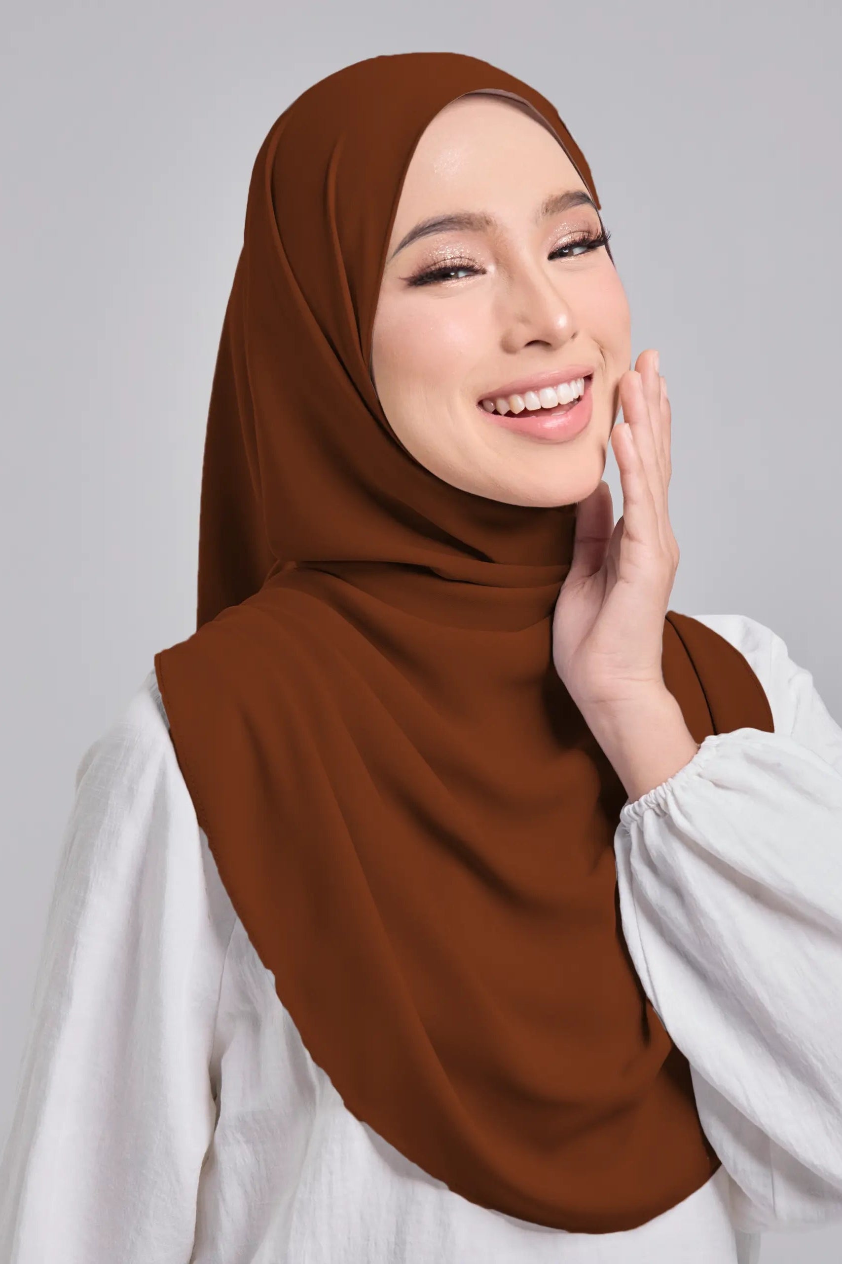 Woman wearing a brown hijab and white top against a gray background