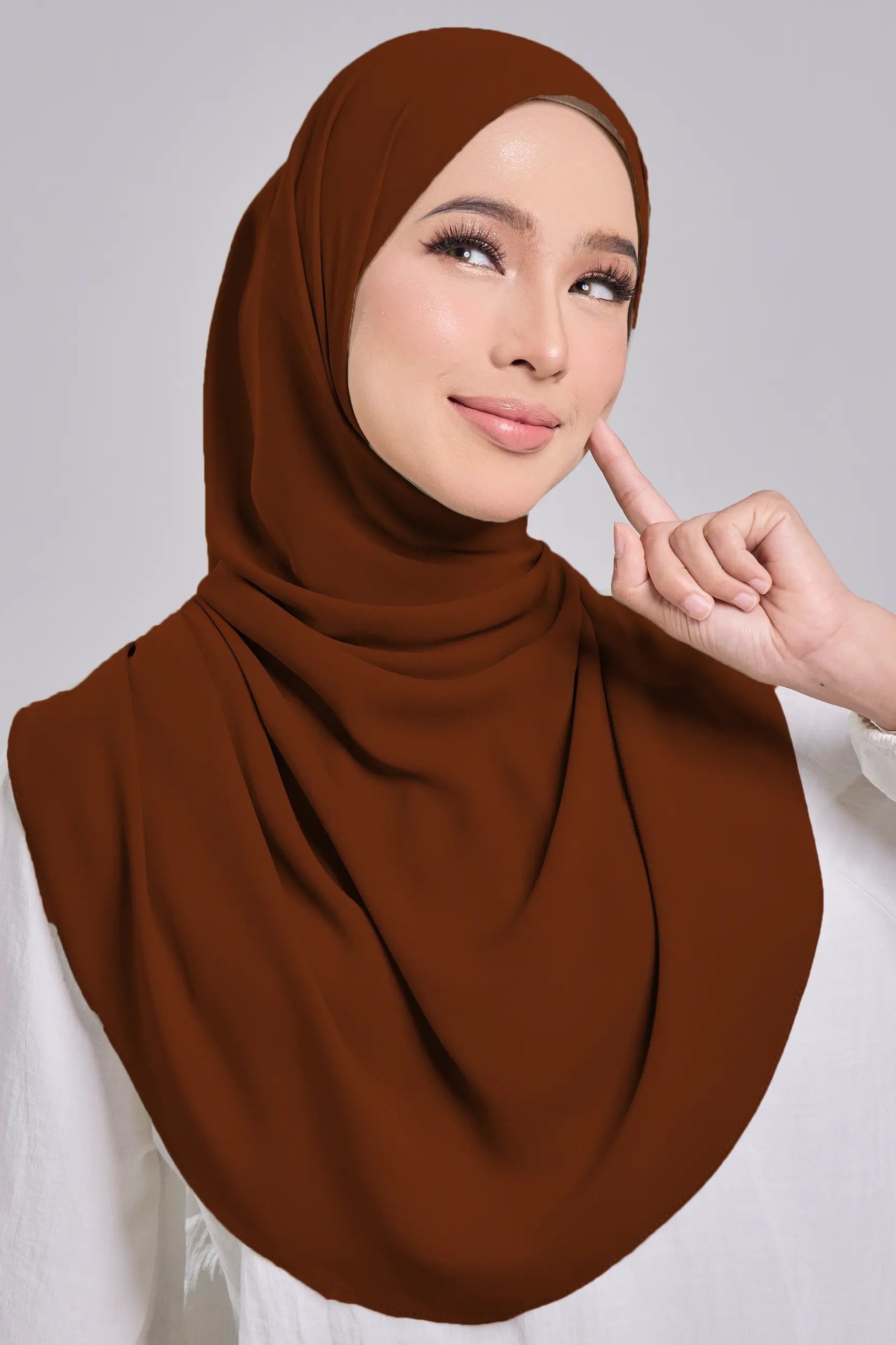 Woman wearing a brown hijab and white top against a gray background