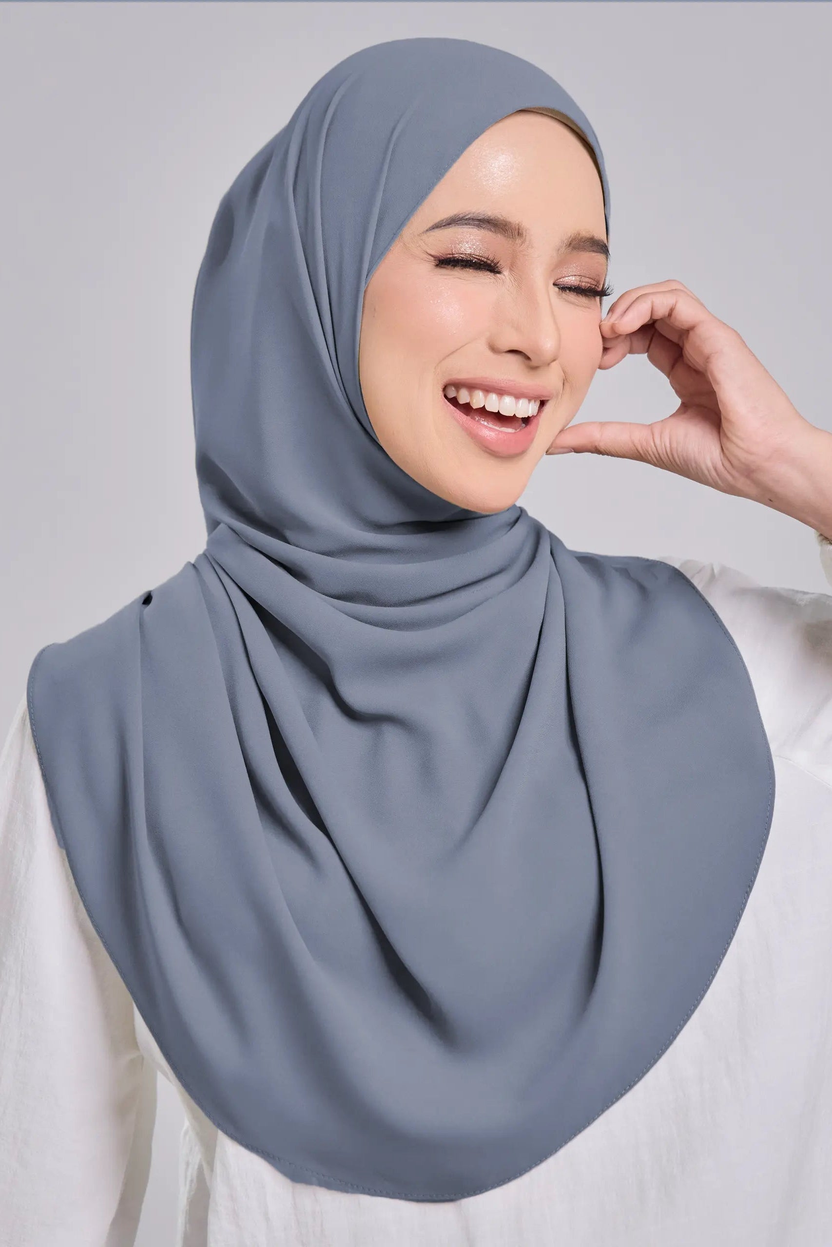 A woman wearing a draped dusty blue-gray hijab and a white long-sleeve top, smiling against a light gray studio background.