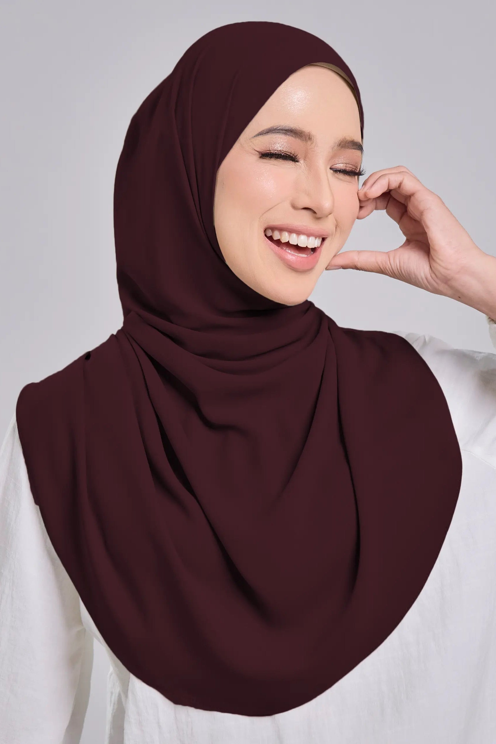 Woman wearing a maroon hijab and white shirt on a gray background