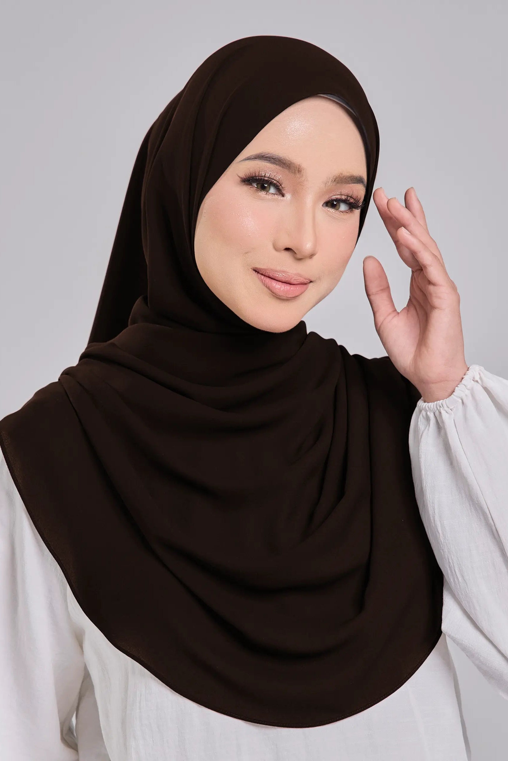 Woman wearing a brown hijab and white blouse against a gray background