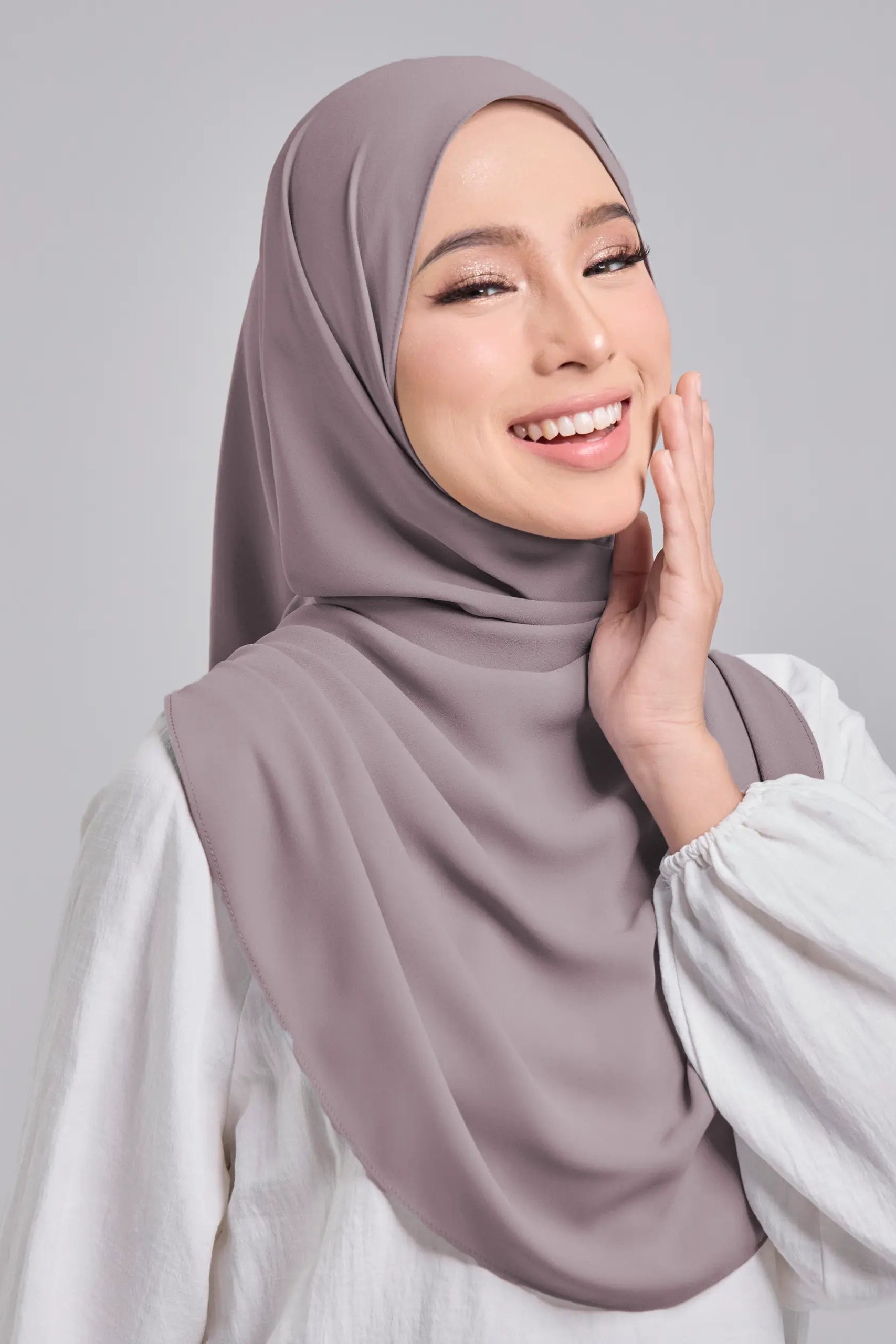 Woman wearing a light brown hijab and white top on a gray background