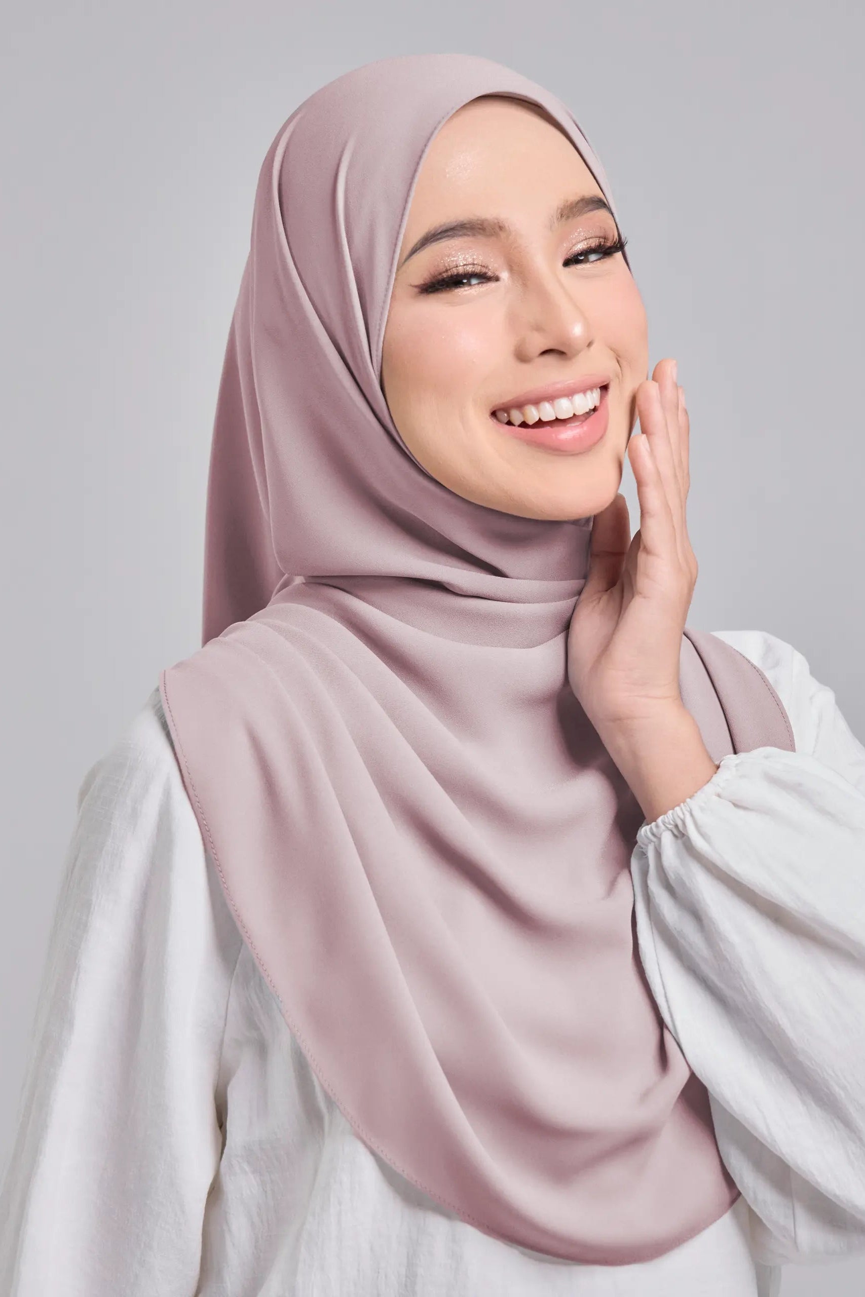 Woman wearing a light pink hijab and white top against a gray background