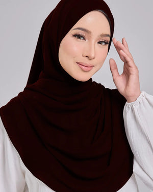 Woman wearing a dark brown hijab and white blouse against a gray background