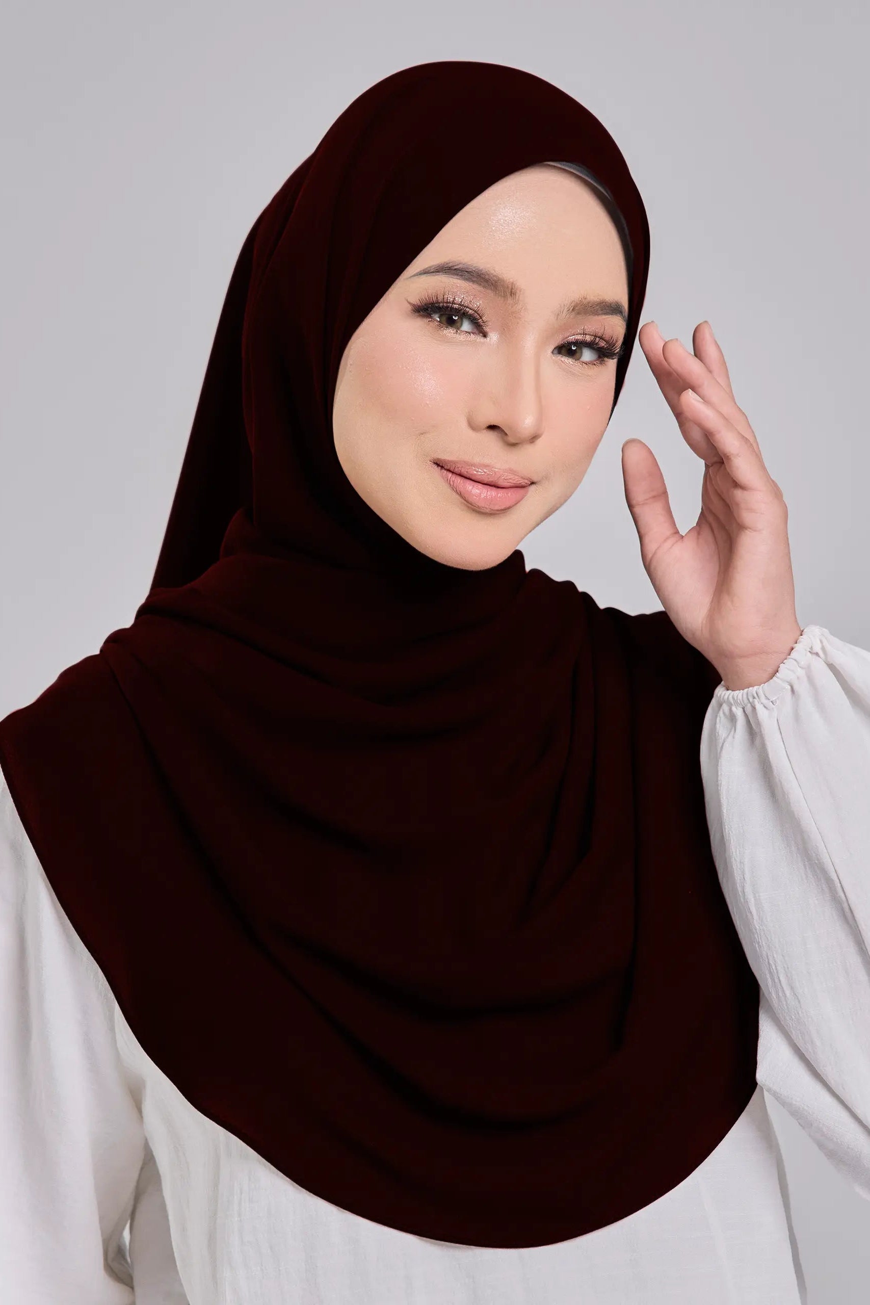 Woman wearing a dark brown hijab and white blouse against a gray background