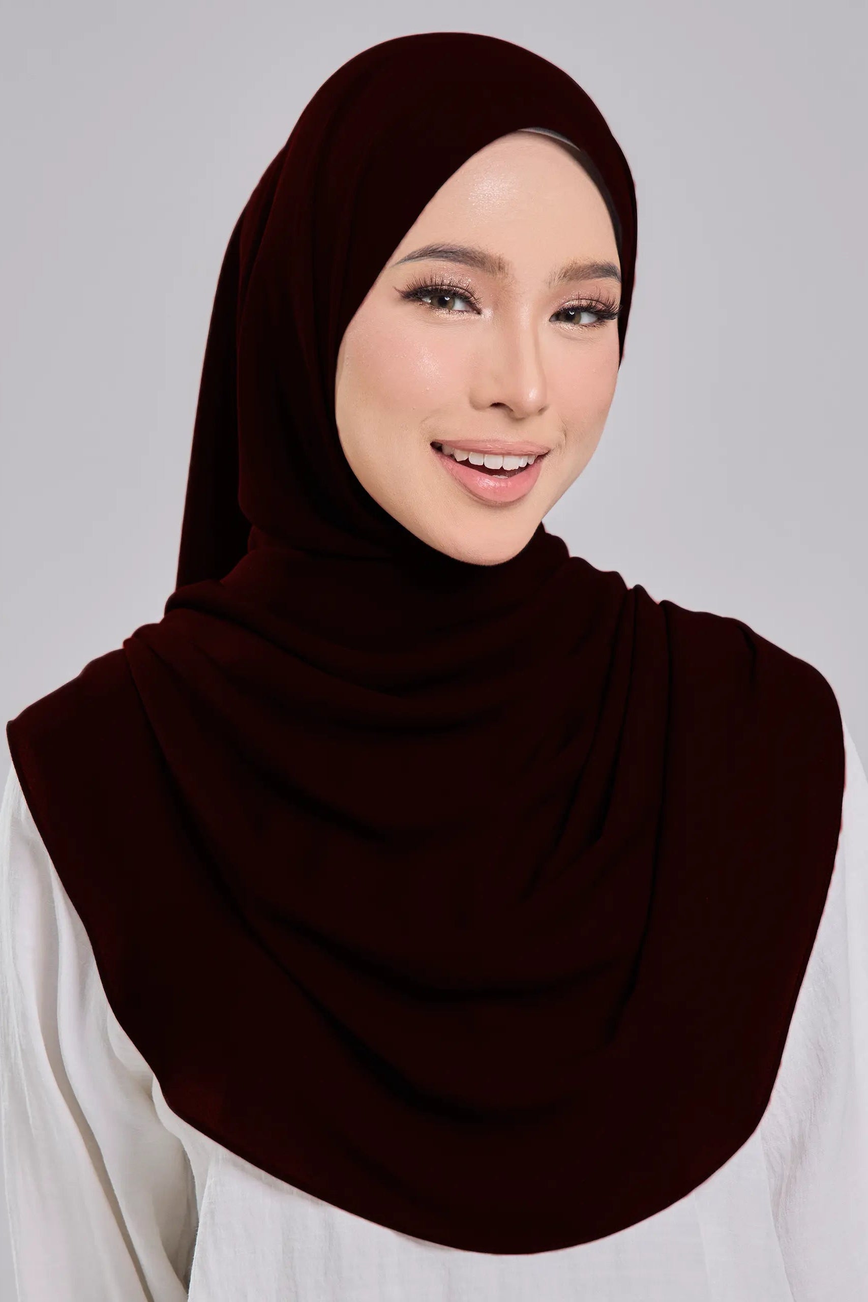 Woman wearing a dark brown hijab and white top on a gray background