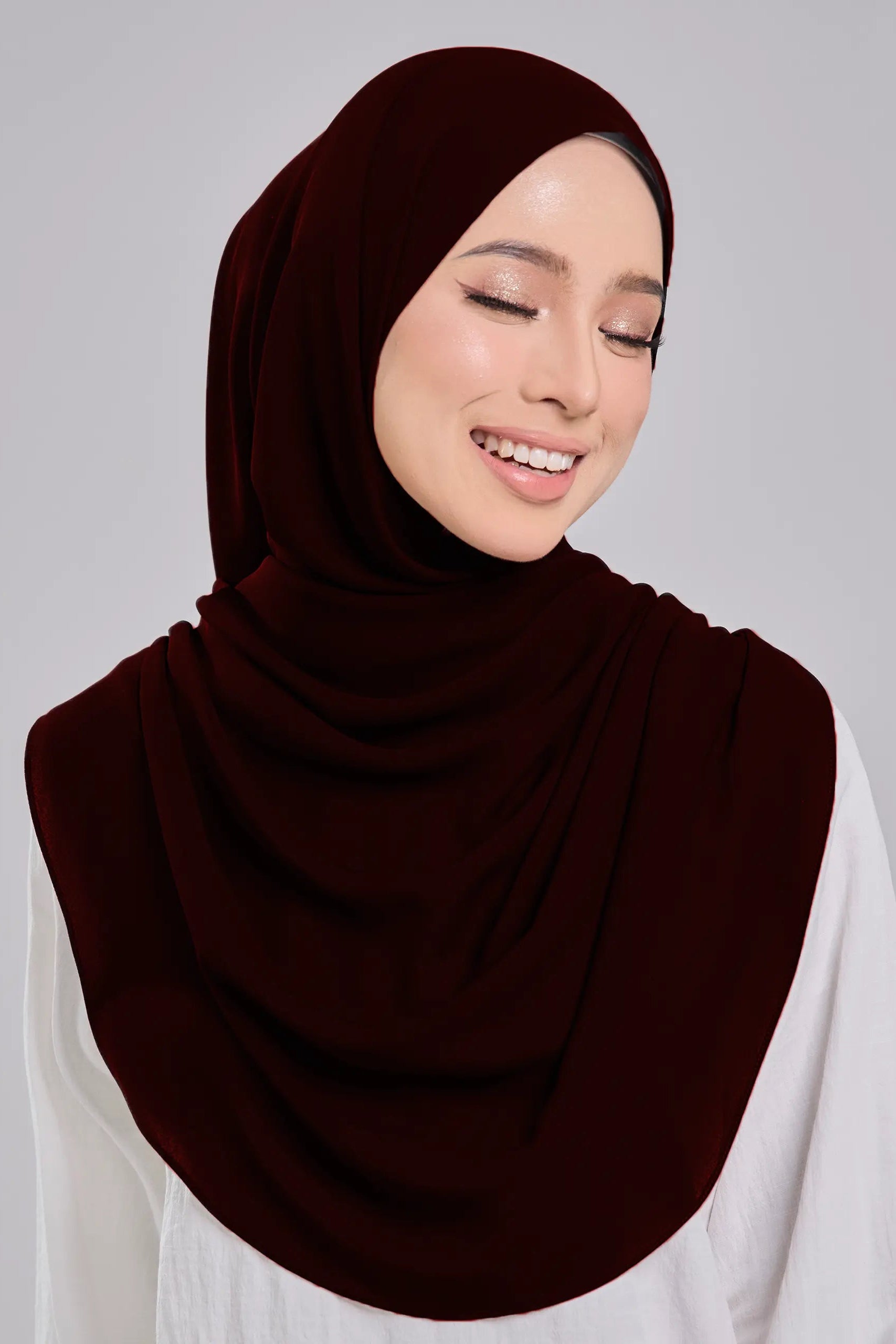 Woman wearing a dark brown hijab and white shirt on a gray background