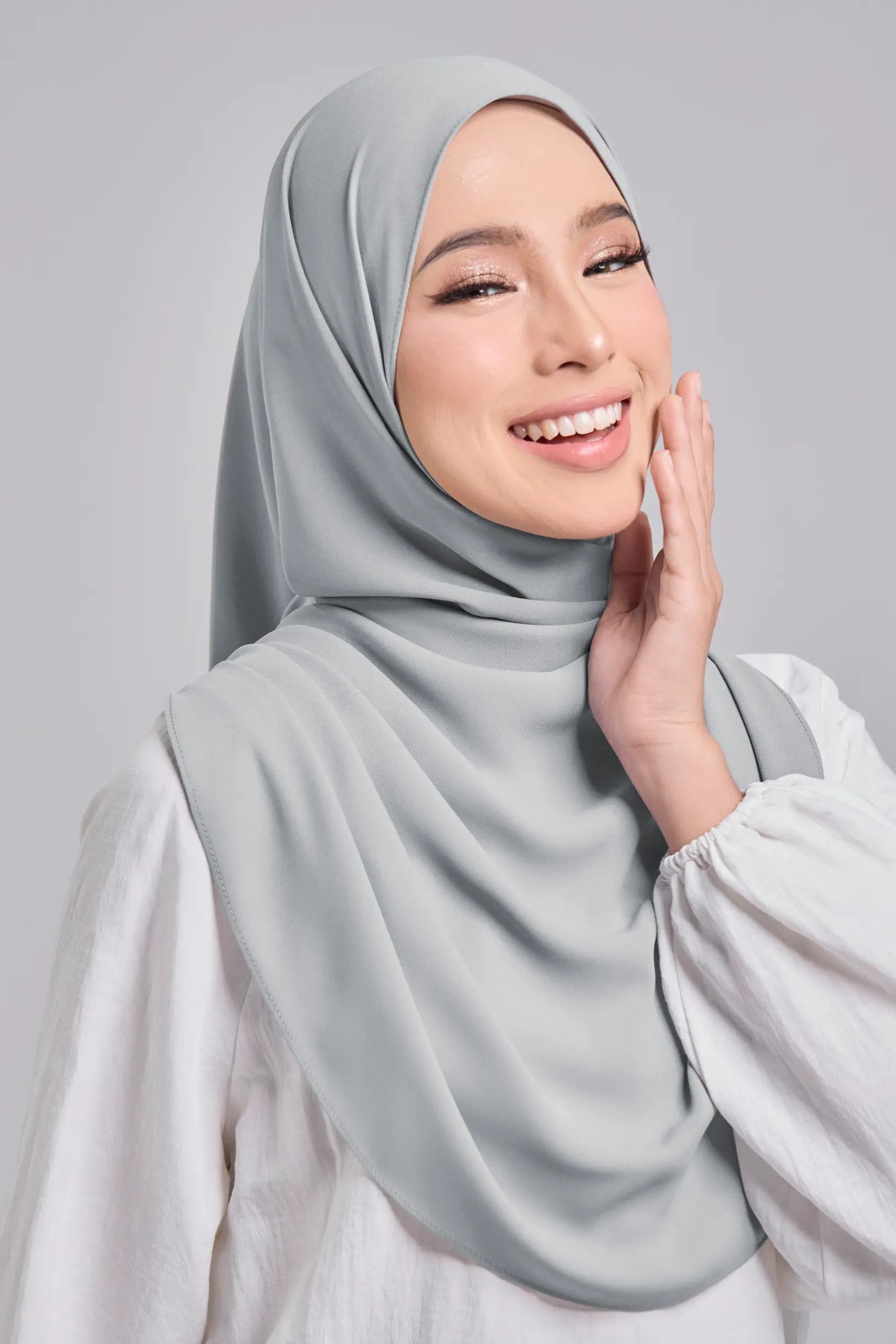 Woman wearing a light gray hijab and white shirt on a gray background