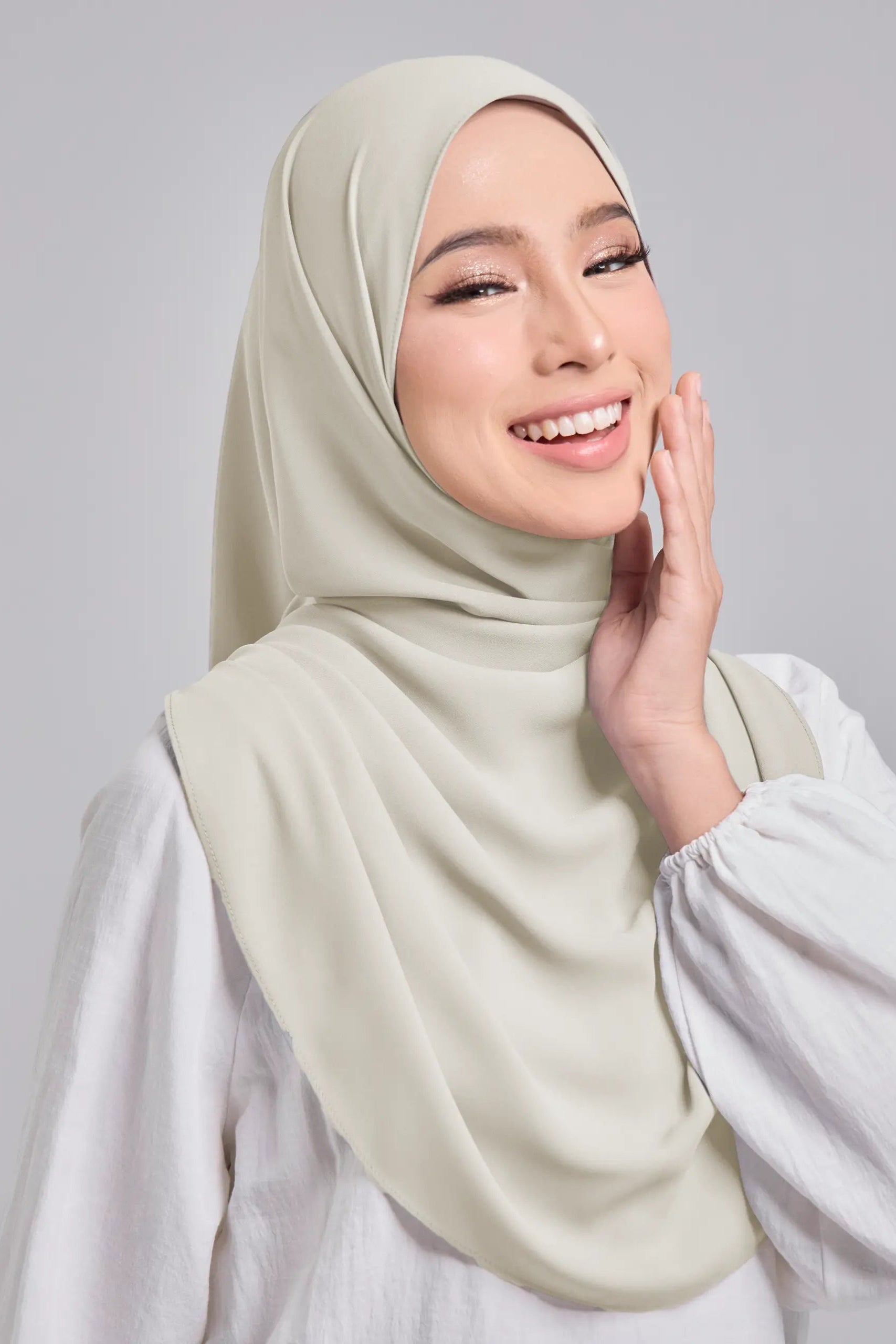 Woman wearing a light-colored hijab and white top against a plain background