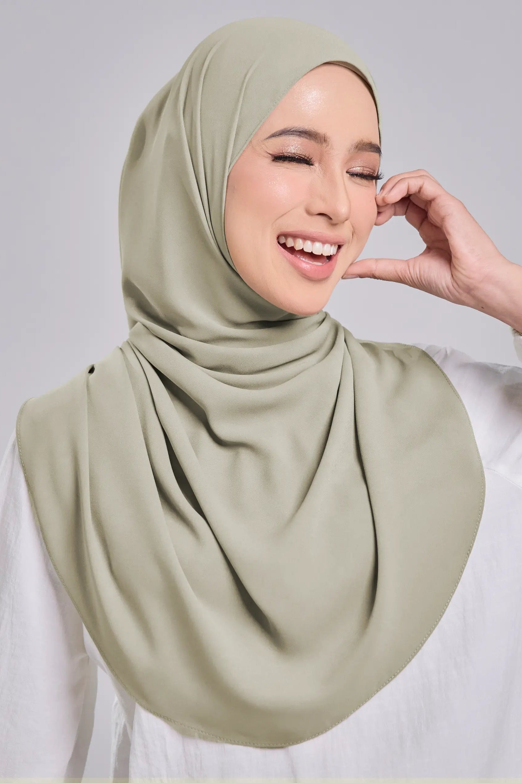 A smiling woman wearing a draped sage green hijab and a white long-sleeve top, posed against a light gray studio background.