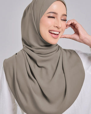 A woman wearing a draped taupe-gray hijab and a white long-sleeve top, smiling against a solid light gray background.