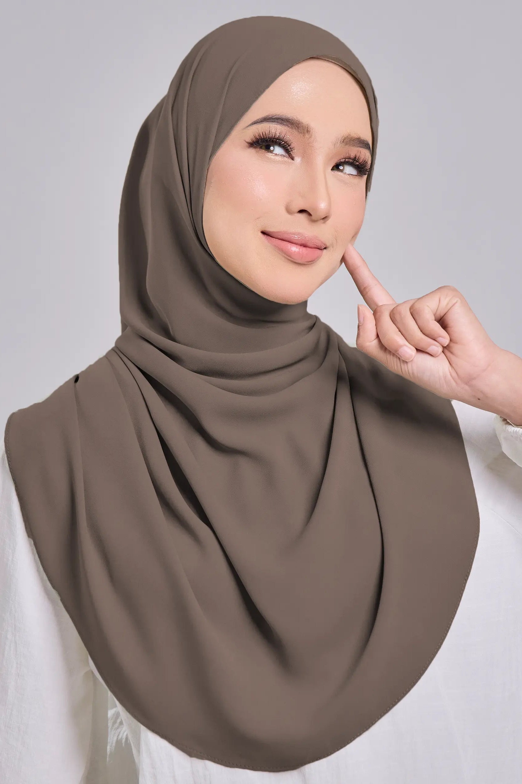 Woman wearing a brown hijab and white top against a gray background