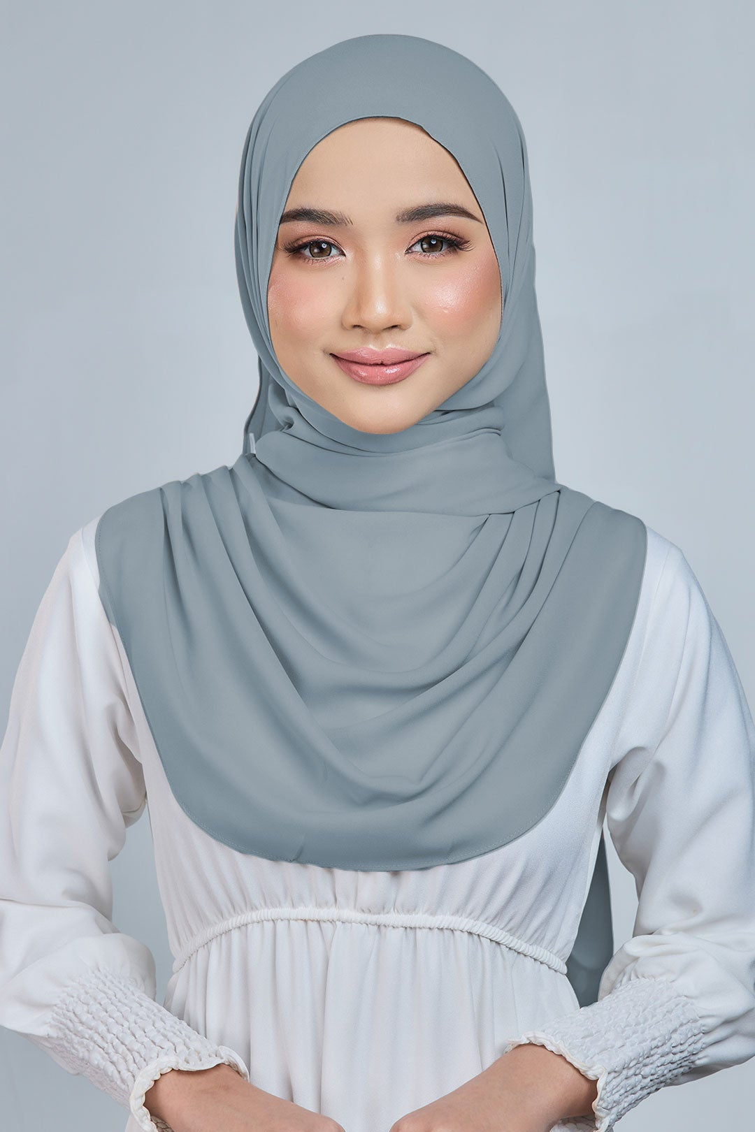 Woman wearing a light gray hijab and white dress against a gray background