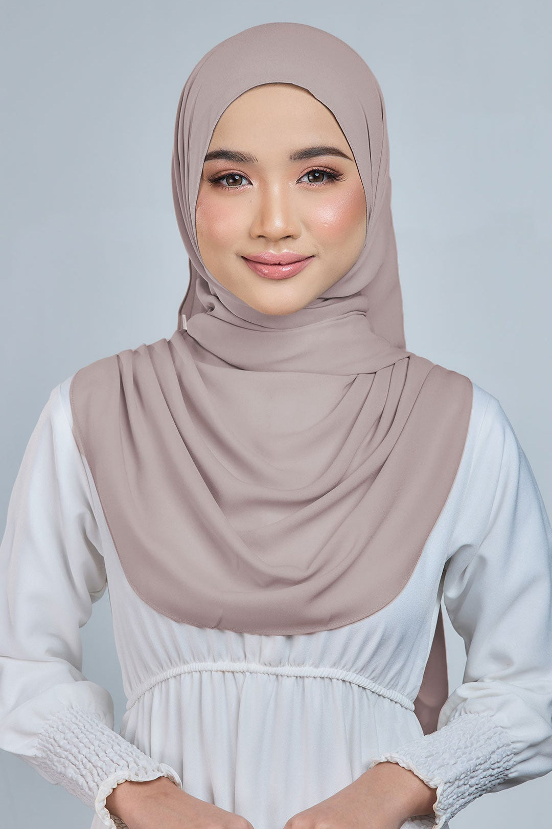 Woman wearing a beige hijab and white dress against a gray background