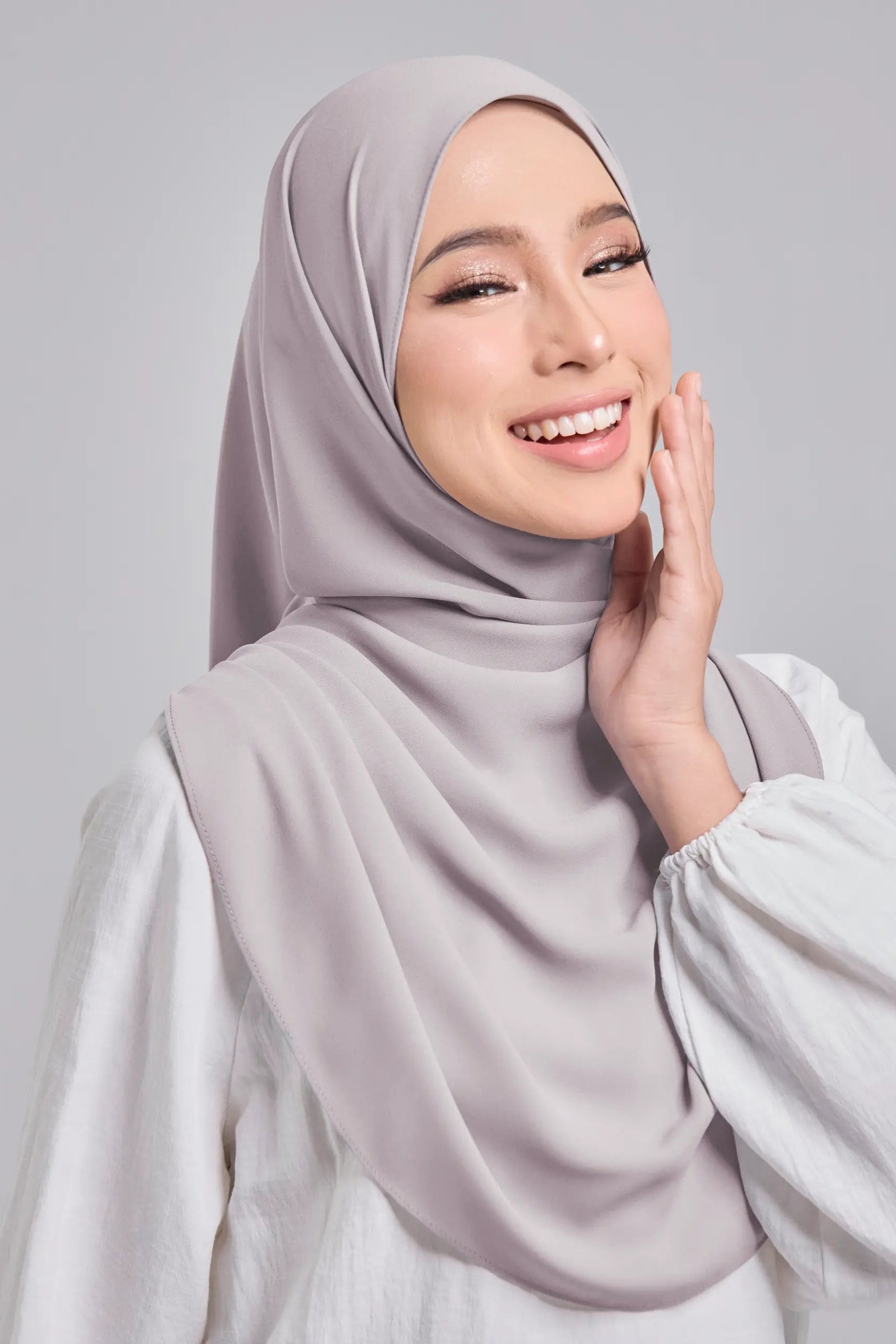 Woman wearing a light gray hijab and white top against a plain background