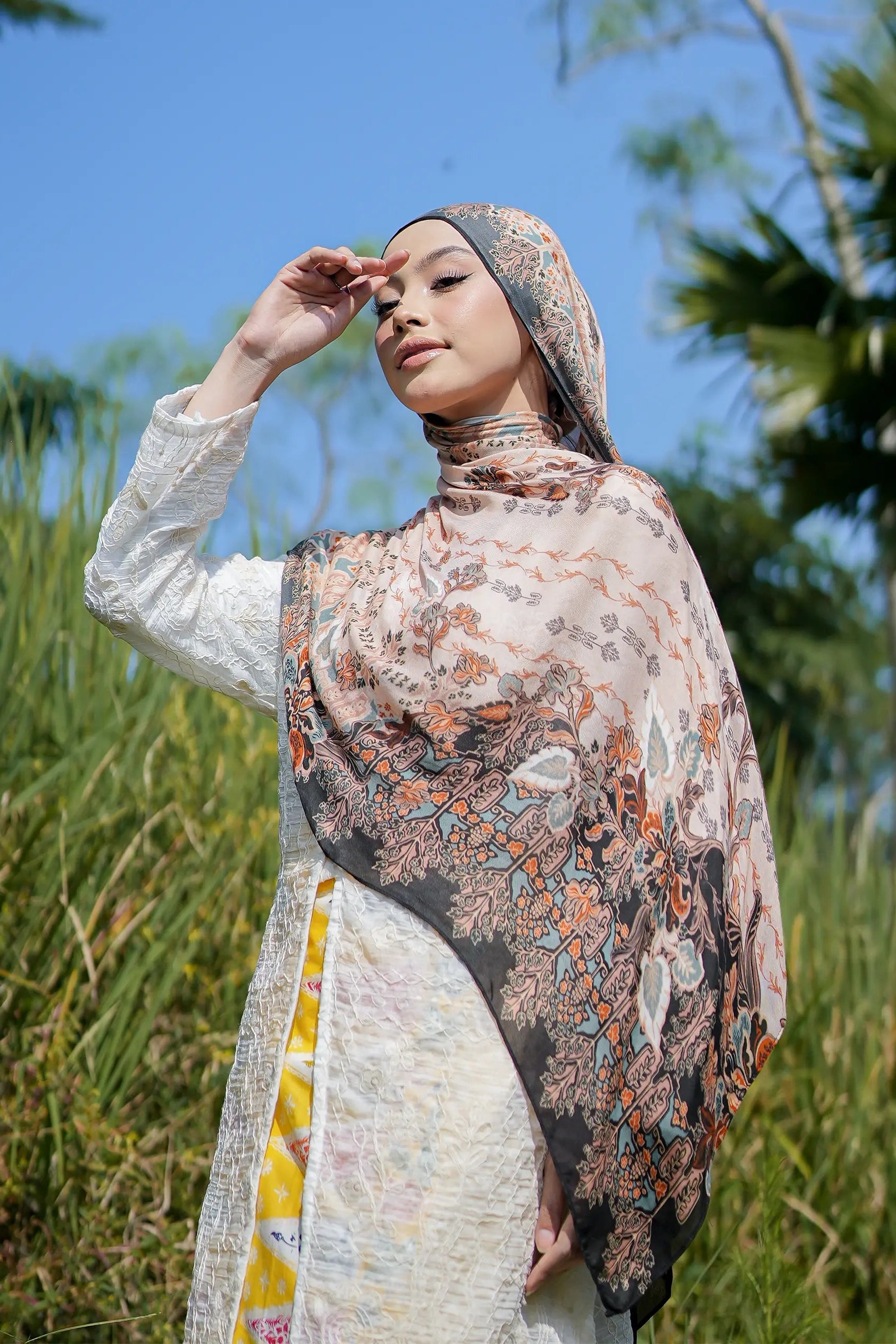 Woman wearing a patterned hijab and dress in a natural setting with trees and blue sky.