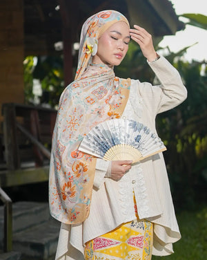 Woman in traditional outfit holding a fan outdoors with greenery around