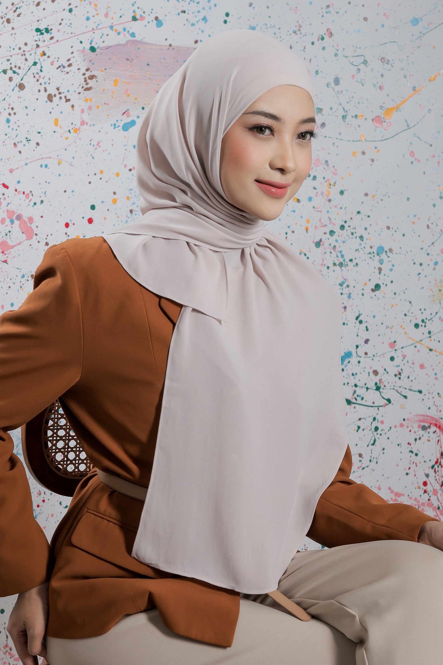 Woman wearing a beige hijab and brown top sitting against a colorful abstract background