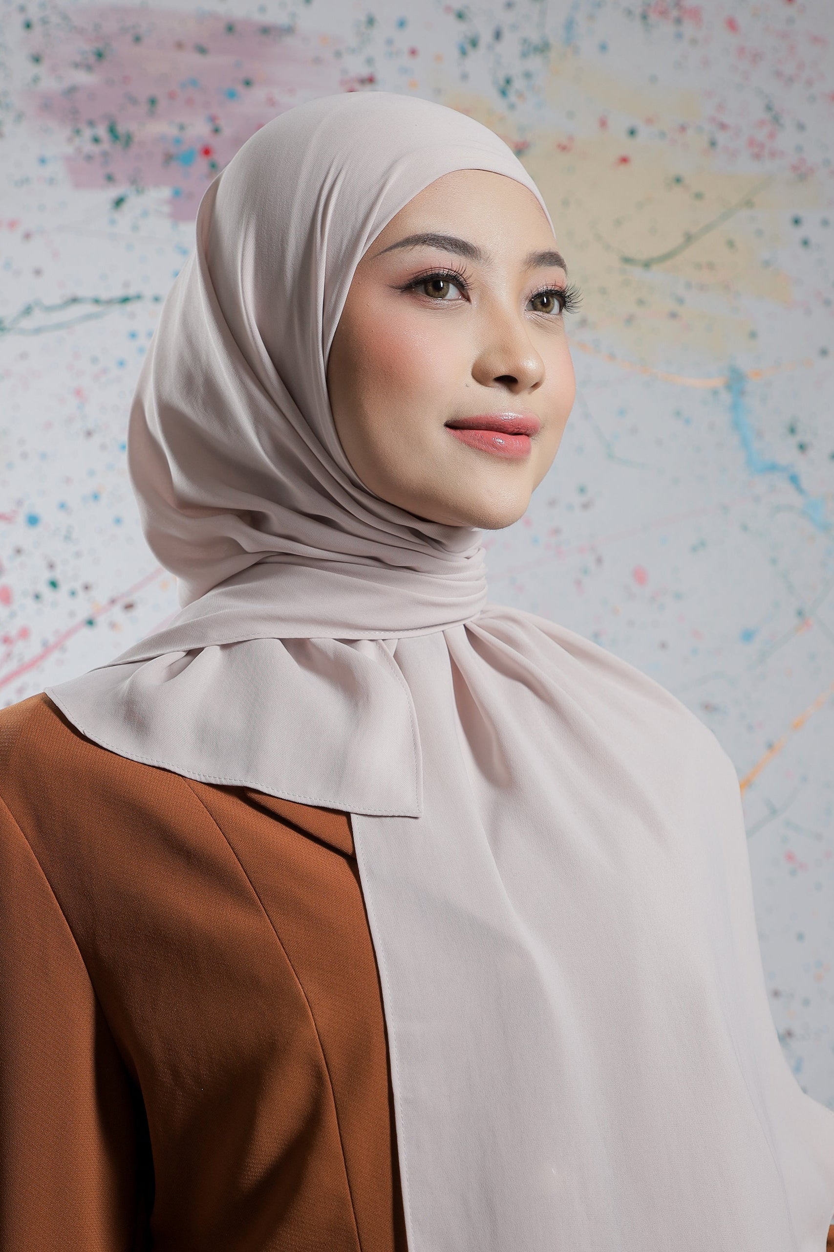 Woman wearing a beige hijab and brown top against a colorful abstract background