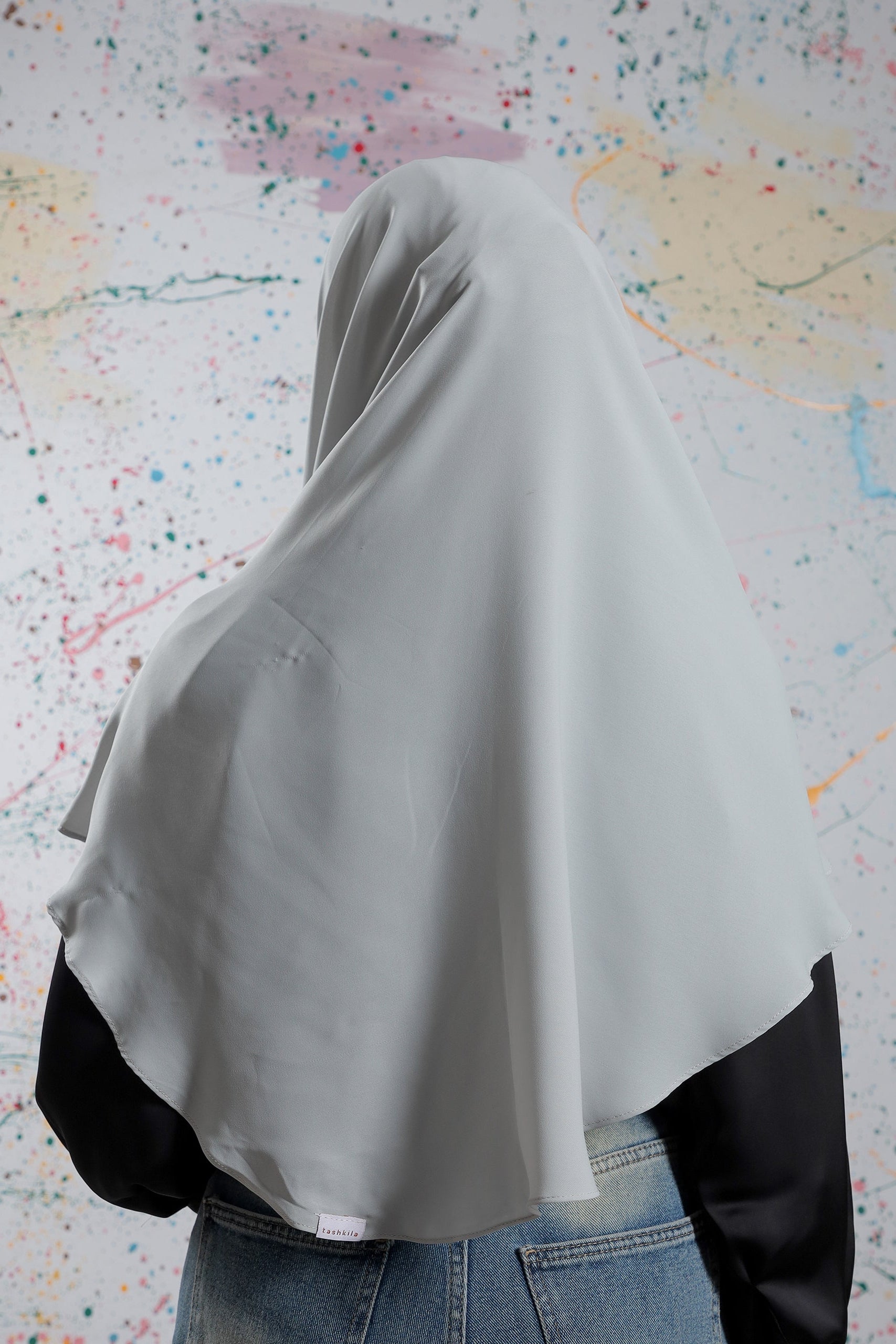 Person wearing a light gray hijab with black sleeves against a colorful abstract background