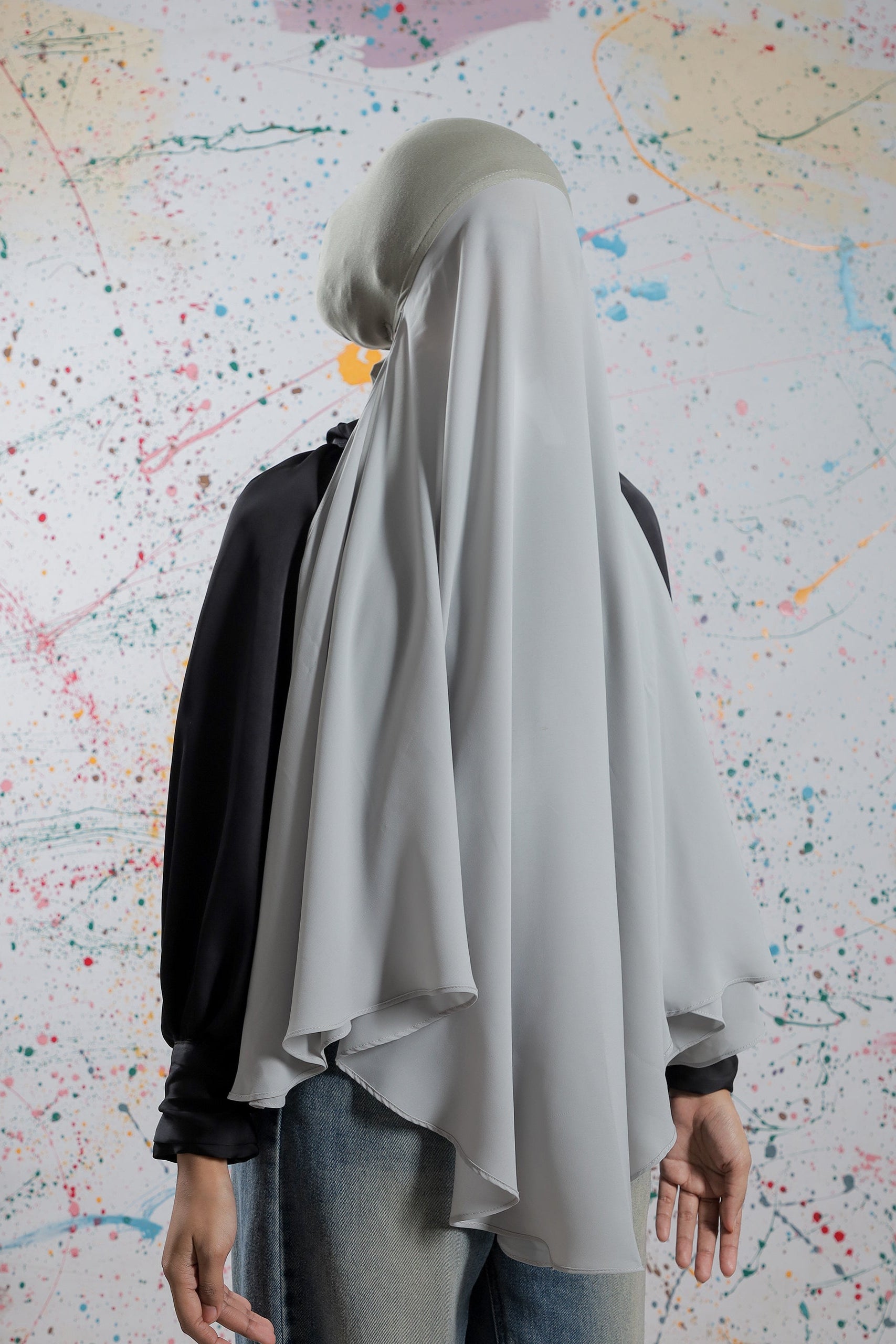 Person wearing a light gray hijab in front of a colorful abstract wall.