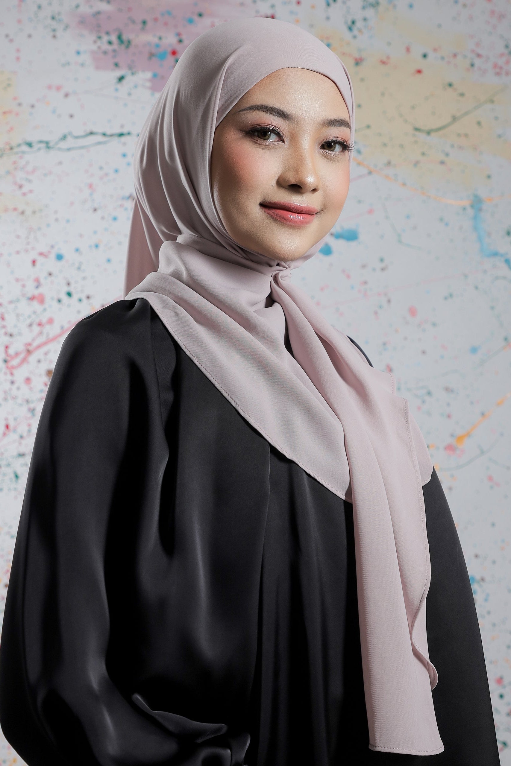 Woman wearing a light pink hijab and black outfit against a colorful abstract background