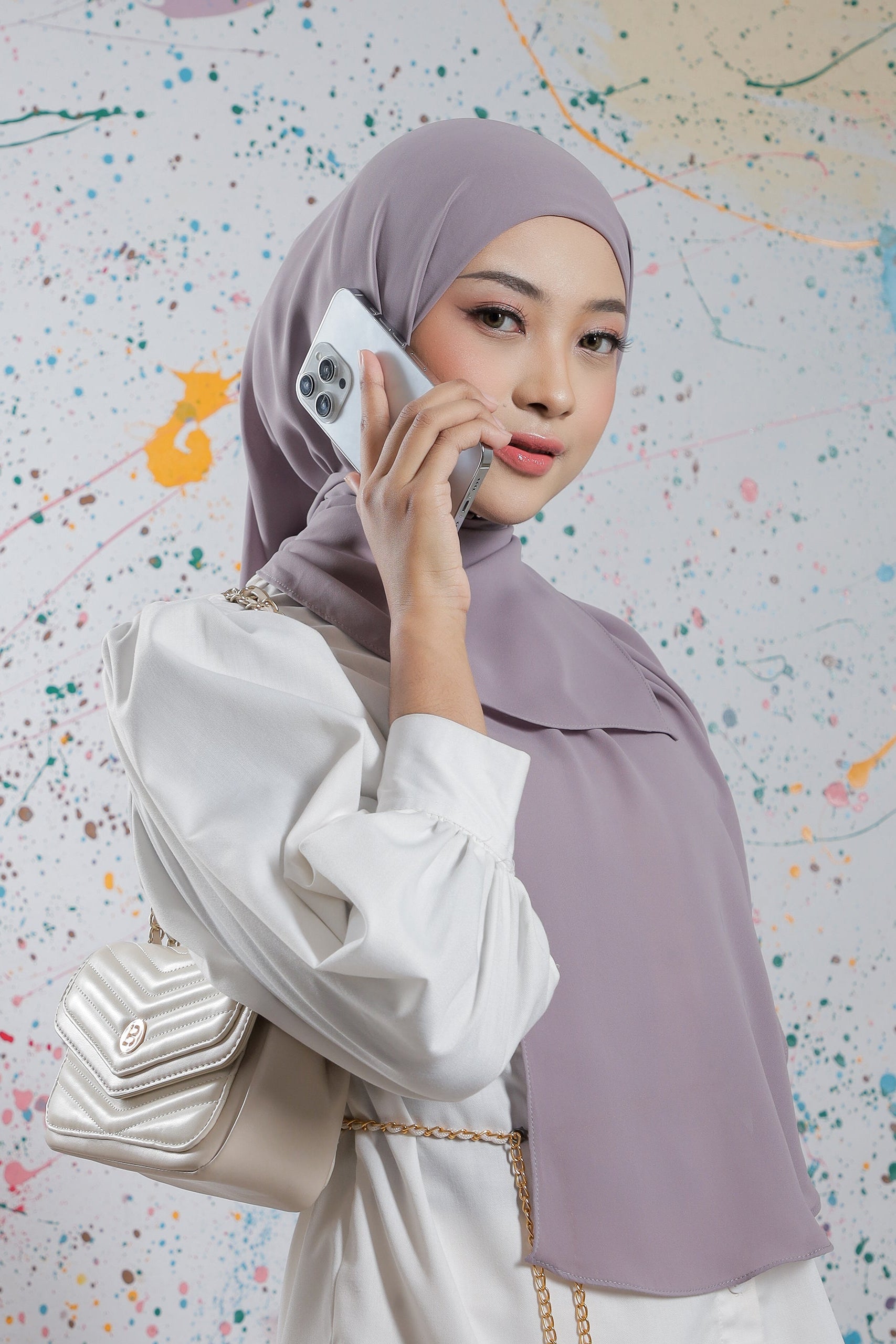 Woman wearing a light purple hijab and white outfit, holding a phone with a colorful abstract background.