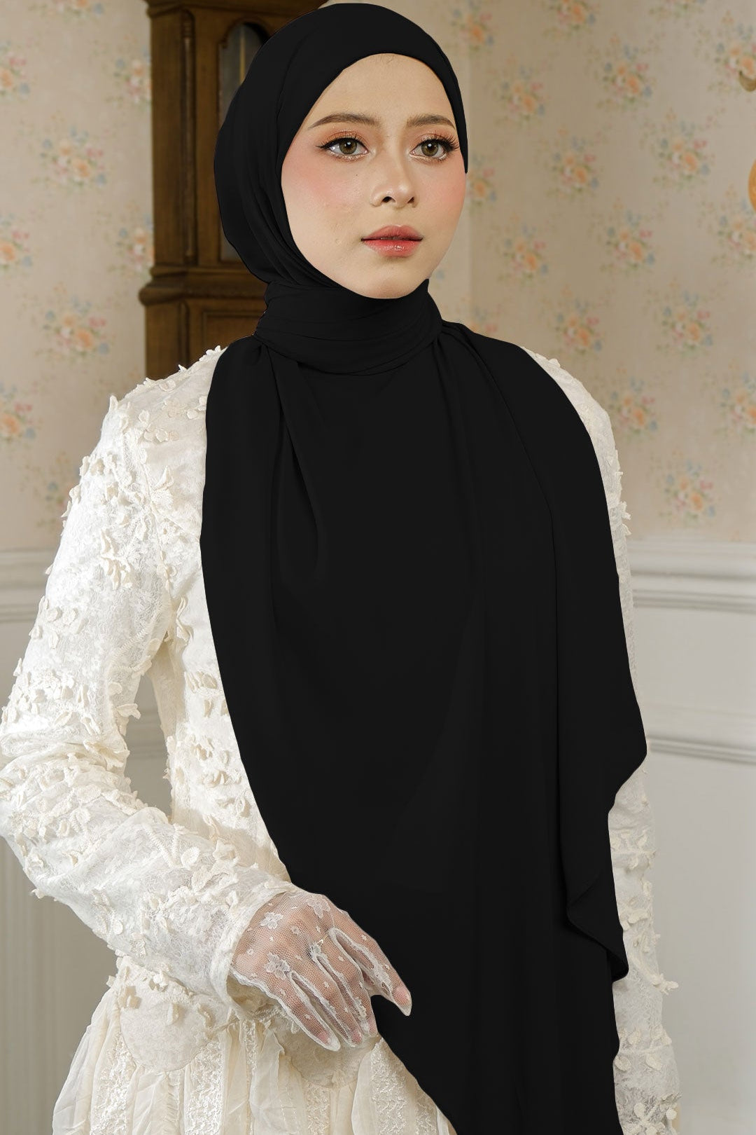 Woman wearing a black hijab and white lace dress in a decorated room.