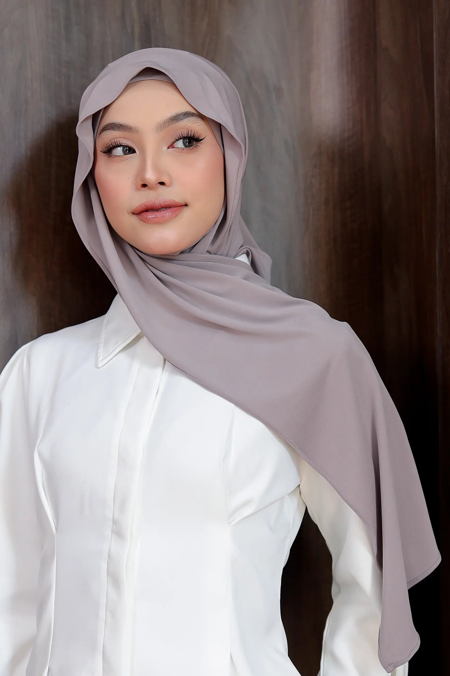 Woman wearing a light gray hijab and white shirt against a brown background