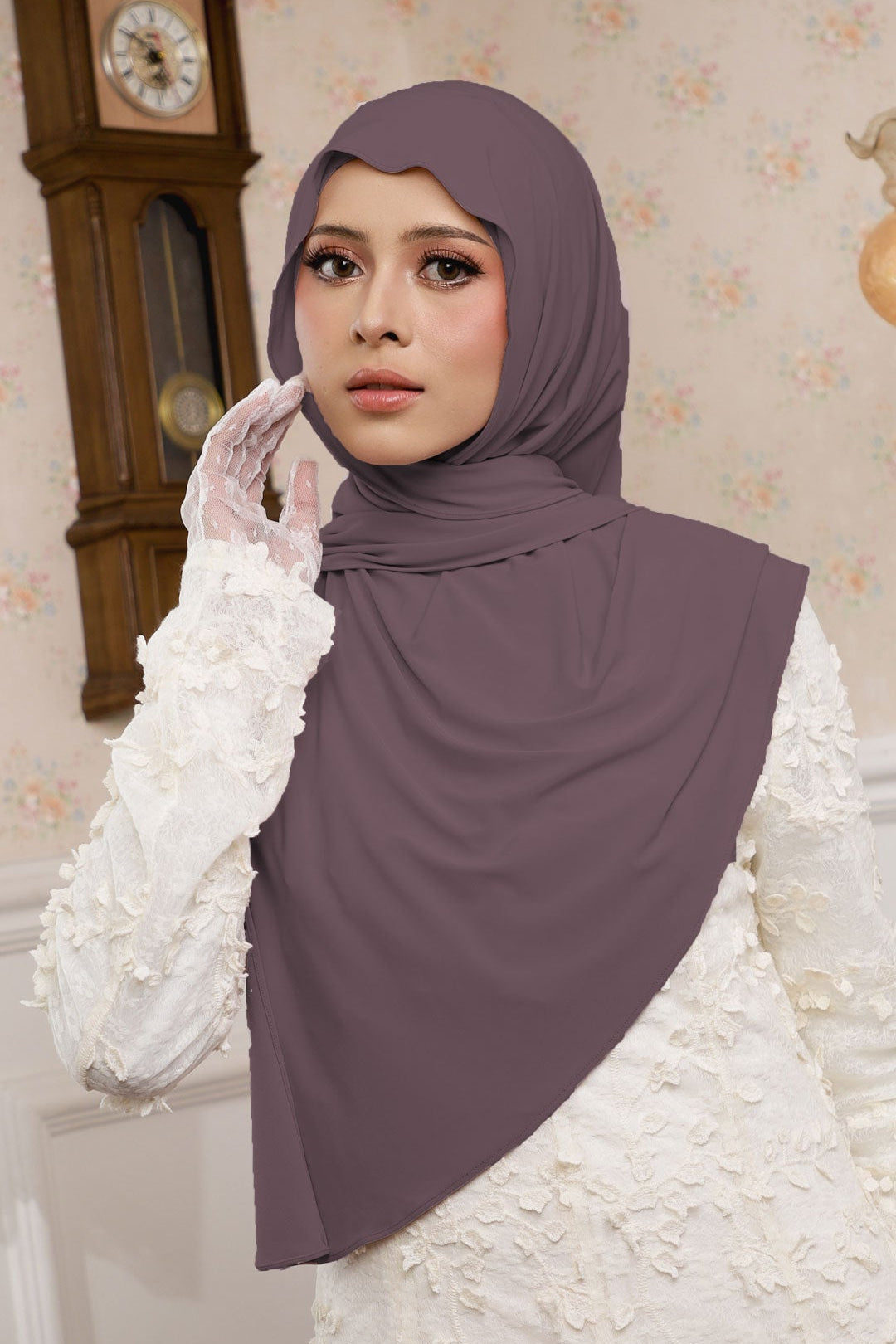 Woman wearing a purple hijab and white lace garment in a decorated room.