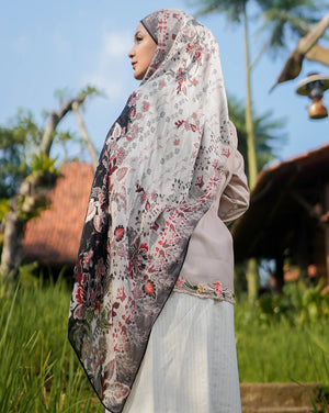 Woman wearing a floral hijab in a natural setting with trees and traditional architecture.