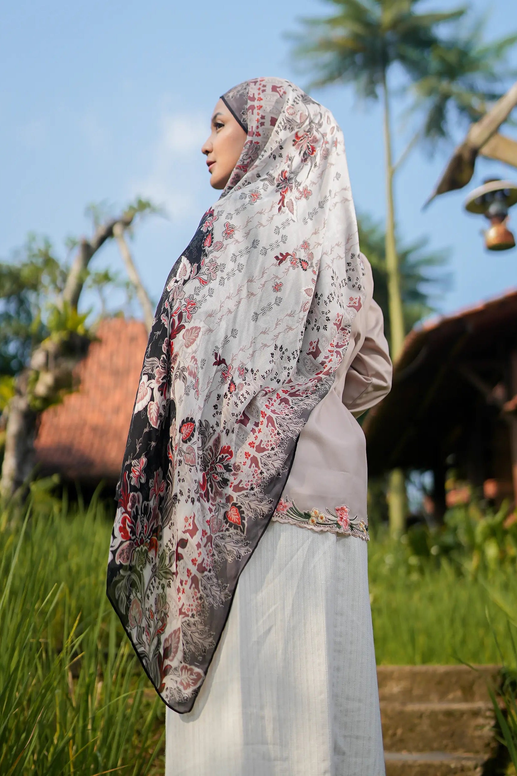 Woman wearing a floral hijab in a natural setting with trees and traditional architecture.