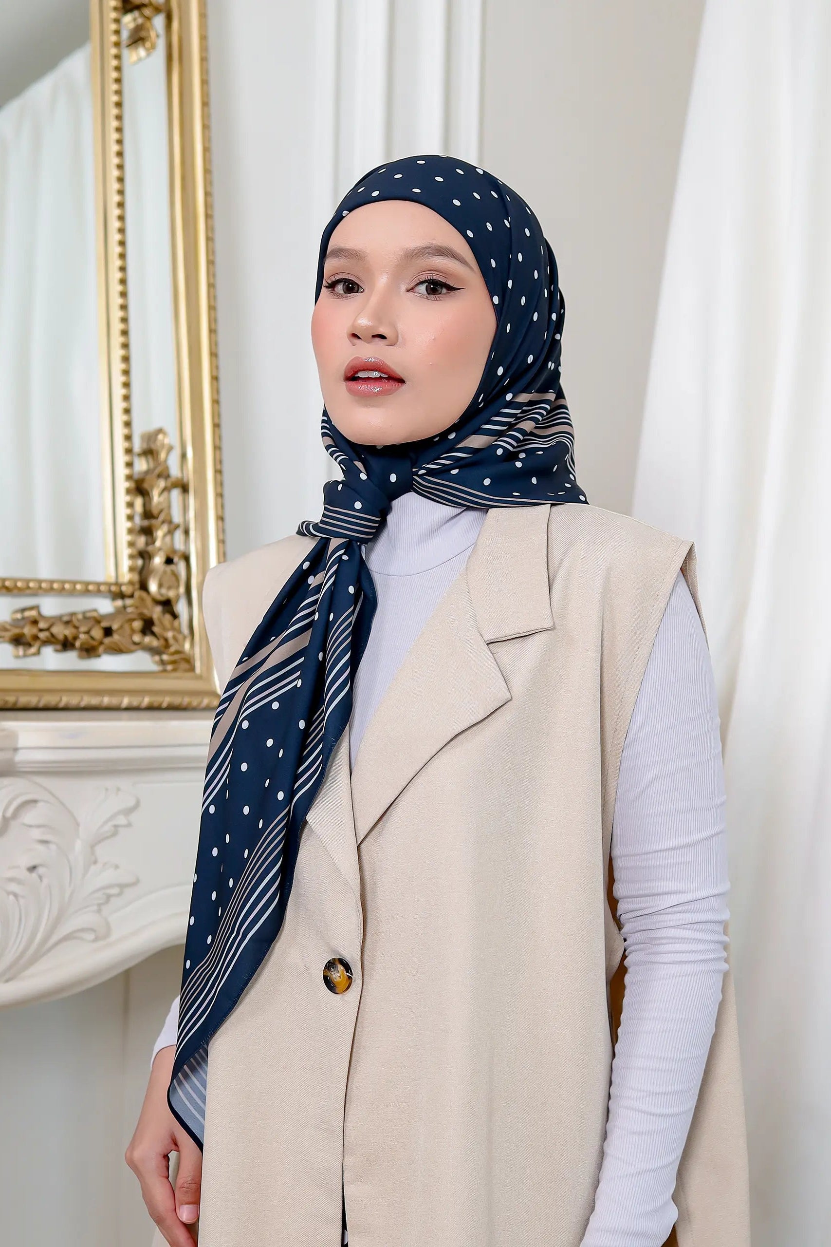 Woman wearing a navy polka dot hijab with a matching scarf in a room with white curtains and a decorative mirror.