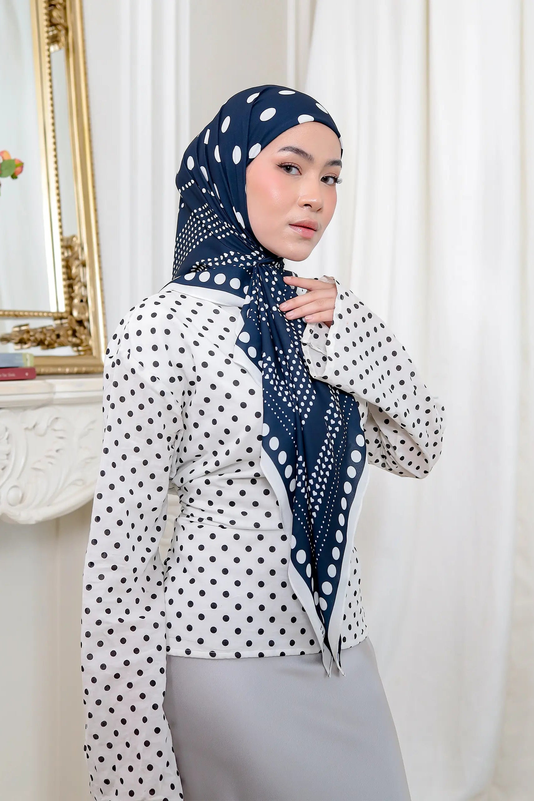 Woman wearing a white blouse with black polka dots and a navy hijab with white patterns, standing in a room with decorative elements.