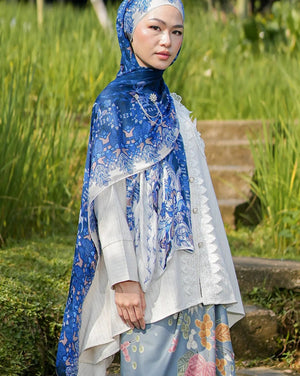 Woman wearing a blue and white floral hijab and dress standing in a natural setting with greenery.