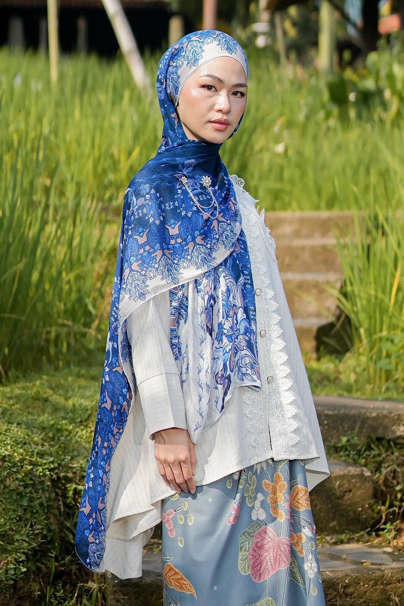 Woman wearing a blue and white floral hijab and dress standing in a natural setting with greenery.