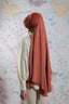 Person wearing a long, rust-colored hijab in front of a colorful abstract wall.